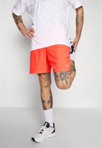 Mann med tatoveringer som strekker ut beinet, iført hvite New Balance-sko, hvite sokker, klare oransje shorts, og en hvit t-skjorte mot en nøytral bakgrunn.