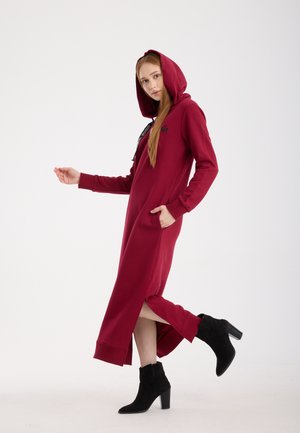 Lange bordeauxrode sweatjurk met capuchon, zijslitsen, voorzakken en zwarte accenten, gecombineerd met zwarte enkellaarsjes met hak.