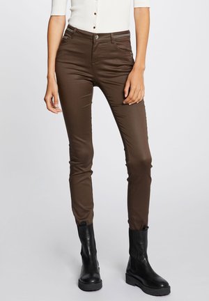 Mujer con pantalones ajustados marrones, camiseta blanca acanalada de manga corta con botones y botines negros, de pie frente a un fondo liso.