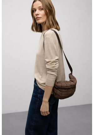 Frau mit hellbraunen Haaren, die einen beigen Pullover und dunkle Jeans trägt, mit einer braunen gesteppte Schultertasche vor einem schlichten grauen Hintergrund.