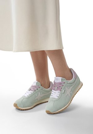 Hellgrüne Wildleder-Sneaker mit pinken Schnürsenkeln und weißer Zunge, getragen von einer Person in einem cremefarbenen Rock, stehend auf weißem Boden.