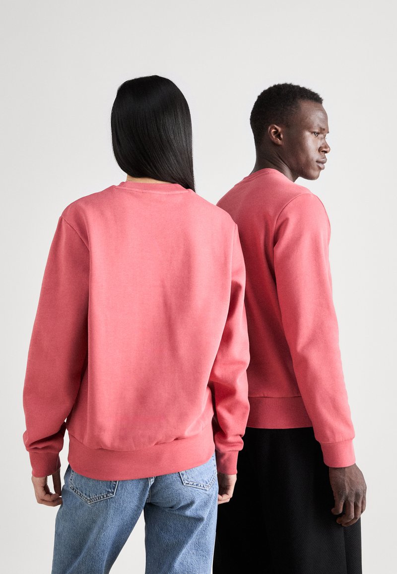 Dos individuos llevan sudaderas de pullover a juego en un color coral sólido. Las sudaderas tienen mangas largas y puños acanalados.