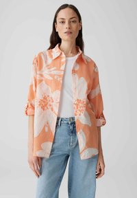 Camicia con motivo floreale in corallo arancione con fiori bianchi, tessuto leggero, maniche lunghe arrotolate, vestibilità comoda, chiusura anteriore con bottoni, abbinata a jeans chiari.