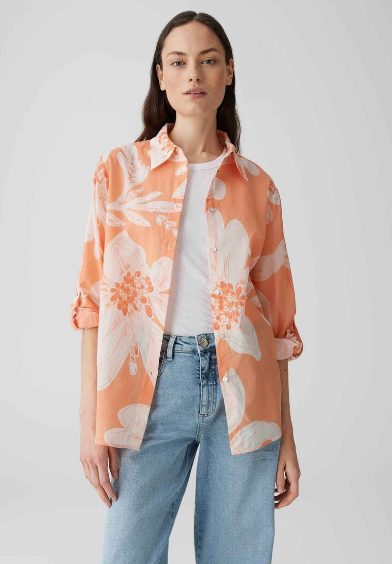 Camicia con motivo floreale in corallo arancione con fiori bianchi, tessuto leggero, maniche lunghe arrotolate, vestibilità comoda, chiusura anteriore con bottoni, abbinata a jeans chiari.