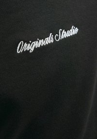Fekete szövet, sima textúrával, fehér hímzett "Originals Studio" felirattal, kézzel írt betűtípussal.