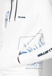 Sudadera blanca con capucha que presenta diseños gráficos en azul, incluyendo formas geométricas y líneas. Texto del logo en azul en el pecho y la manga.