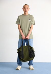 Sac de sport vert olive avec des accents noirs, forme structurée, doubles poignées, poches latérales en maille et fermeture à cordon réglable.