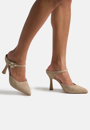 Beige Wildleder High-Heel-Mules mit spitzem Zehenbereich. Mit einem dünnen Knöchelriemen mit dekorativen Verzierungen und einem skulpturierten Absatz.