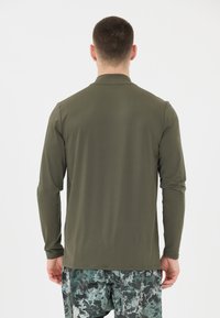 Langärmliges, olivgrünes Hemd aus glattem Stoff, mit Kragen und geradem Schnitt. Getragen mit gemusterten, dunkelgrünen Shorts.