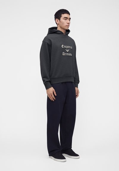 Jeune homme debout portant un sweat à capuche Emporio Armani gris foncé, un pantalon noir ample et des baskets noires à semelles blanches sur un fond uni.