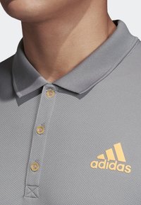 Šedé polo tričko vyrobené z texturovaného materiálu s tříknoflíkovou légou. Zlaté logo Adidas na levé straně.