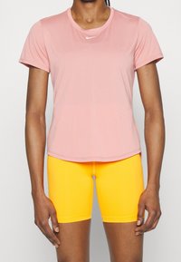 Camiseta deportiva rosa de manga corta con cuello redondo y logo blanco, combinada con pantalones cortos amarillos ajustados que llegan hasta la mitad del muslo.