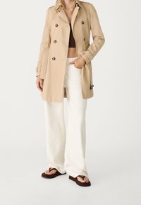 Beige trenchcoat med dubbelknäppt design, stora knappar och axelklaffar, kombinerad med vita vida byxor och bruna flip-flops.