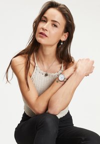 Zilveren horloge met een ronde wijzerplaat, gecombineerd met een gelaagde ketting en oorbellen. Het model draagt een lichte gebreide top en zwarte broek.