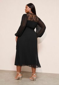 Fekete pliszírozott ruha áttetsző csipkebetétekkel a háton és hosszú ujjakkal. Ezüst pántos magassarkúval viselve. Sima textúra.