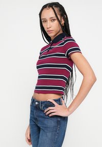 Polo majica kratkih rukava u bordo boji s tamnoplavim i bijelim horizontalnim prugama. Ima ovratnik i skraćeni rub. Nošena s plavim trapericama.