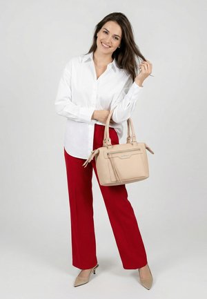 Vrouw die lacht, gekleed in een witte blouse en rode broek, een beige handtas vasthoudend en haar haar aanrakend, staande tegen een eenvoudige lichte achtergrond.