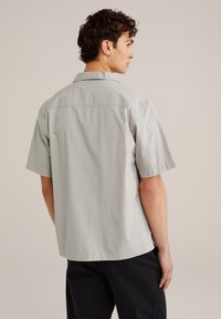 Camisa de manga corta gris claro con un cuello estructurado, textura de tela suave y un dobladillo recto. Costuras visibles en la parte trasera.