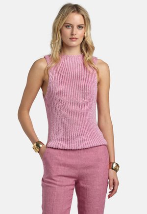 Mujer rubia con top de punto sin mangas rosa y pantalones rosas, de pie con las manos en los bolsillos, llevando amplias pulseras doradas en ambas muñecas.