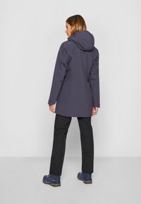 Giacca impermeabile grigia scura con cappuccio, maniche lunghe e orlo leggermente allungato, abbinata a pantaloni neri e scarpe blu.
