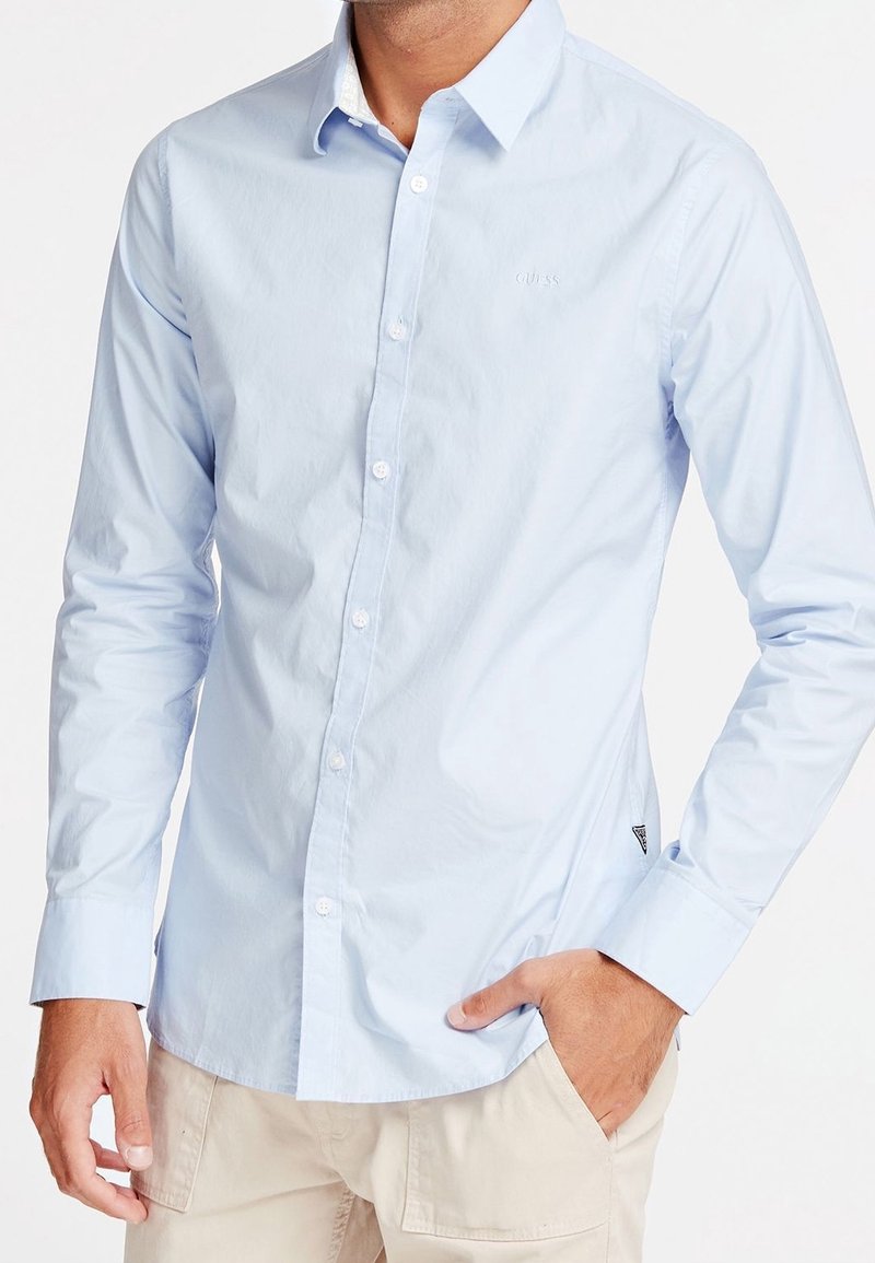 Uomo che indossa una camicia a maniche lunghe blu chiaro con pantaloni beige, una mano in tasca, busto visibile, senza testa mostra.