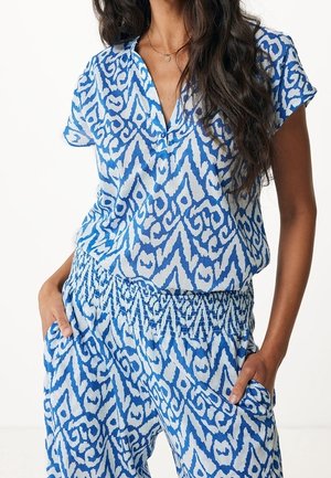 Kobieta ubrana w niebiesko-białą bluzkę z krótkim rękawem w wzory oraz pasujące spodnie z elastycznym pasem, z rękami w kieszeniach, o długich ciemnych włosach.