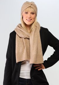 Ensemble écharpe et bandeau en tricot beige, texture douce, design torsadé sur le bandeau, bords côtelés sur l'écharpe. Porté avec un pull noir.