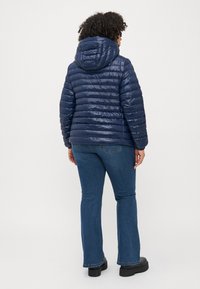 navyblauer gepolsterter Jacket mit Kapuze, ausgestattet mit horizontalen Puffer-Elementen und glänzender Textur, kombiniert mit blauen Schlagjeans und schwarzen Stiefeln.