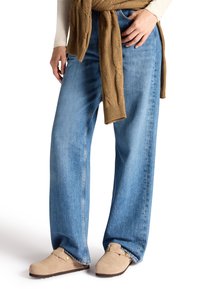 Weite blaue Jeans aus Denim mit einem ausgewaschenen Finish, kombiniert mit einem braunen, kabelgestrickten Pullover, der um die Taille gebunden ist. Helle beige Slipper.