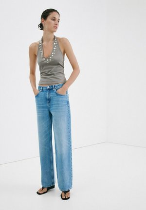 Donna in piedi con jeans a gamba larga blu, top grigio senza maniche a collo alto con dettagli in metallo, mani nelle tasche e indossa tacchi neri open-toe.