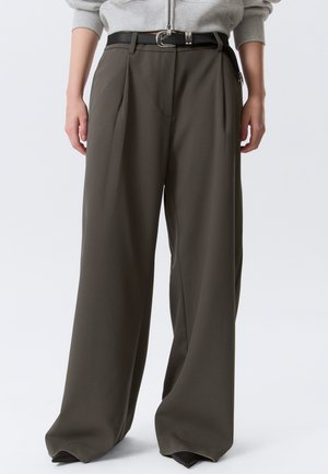 Személy széles szárú olívazöld nadrágban, fekete övvel és világosszürke pulóverben, kezét laza tartásban oldalt.