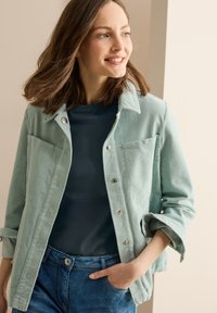 Světle tyrkysová kordurová bunda s límečkem, předními knoflíky a dvěma náprsními kapsami, v kombinaci s námořnickou tričkem a džínovými kalhotami.