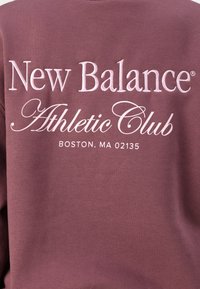Bordo kapuutsärk, millel on helesinise tikandiga tekst: "New Balance Athletic Club" ja allpool tekst "BOSTON, MA 02135". Pehme, tekstuuriga kangas.