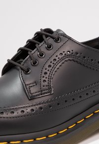 Černá kožená bota s wingtip designem, perforovanými detaily, černými tkaničkami a žlutým kontrastním stehem na podešvi. Viditelná textura gumové podešve.