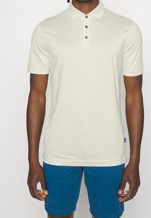 Jasno beżowa koszulka polo z kołnierzykiem, trzema ciemnymi guzikami, krótkimi rękawami i dopasowanym krojem. Połączona z niebieskimi szortami.