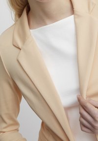 Růžový blejzer s klopovým límcem přes bílý žebrovaný top. Hladká textilní struktura, přiléhavý design a viditelné švy.