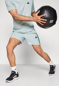 Camisola e calções desportivos azul-claro com detalhes verdes, segurando uma bola medicinal preta. Ténis desportivos pretos e meias brancas com logótipo.