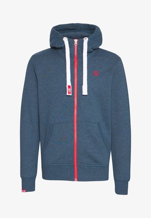 Mann trägt einen marineblauen Kapuzenpullover mit Reißverschluss über einem weißen T-Shirt, schwarze Hose und weiße Sneaker, steht vor einem schlichten Hintergrund.