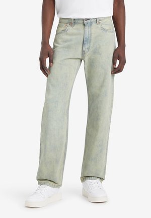 Homme portant un jean droit vert clair délavé, des baskets blanches et un t-shirt blanc, debout les mains détendues le long du corps.