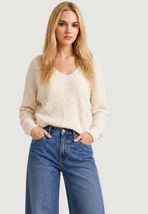 Donna bionda che indossa un maglione di lana color crema e jeans blu a vita alta con le mani in tasca, in piedi davanti a uno sfondo semplice.