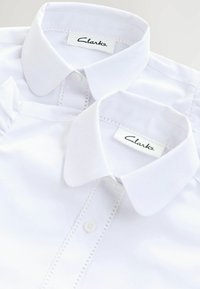 Vita skjortor med klassiska kragar, prydda med knappslå och dekorativt sydda detaljer på sidorna. Varumärkesetikett: Clarks.