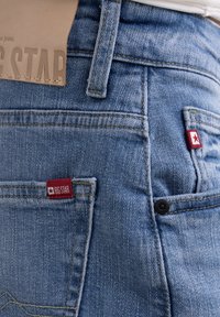 Jean en denim bleu clair avec des coutures visibles, une étiquette en cuir portant la mention "BIG STAR" et de petits accents de logo rouge sur les poches arrière.