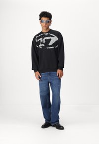 Sort sweatshirt med et stort gråt grafisk design, parret med blå jeans og sorte sko. Model iført sølvfarvede solbriller.