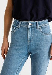 Jeans de tiro alto en azul claro con una textura suave, diseño de cinco bolsillos, cierre de botón plateado y detalles de costura sutiles en las costuras.