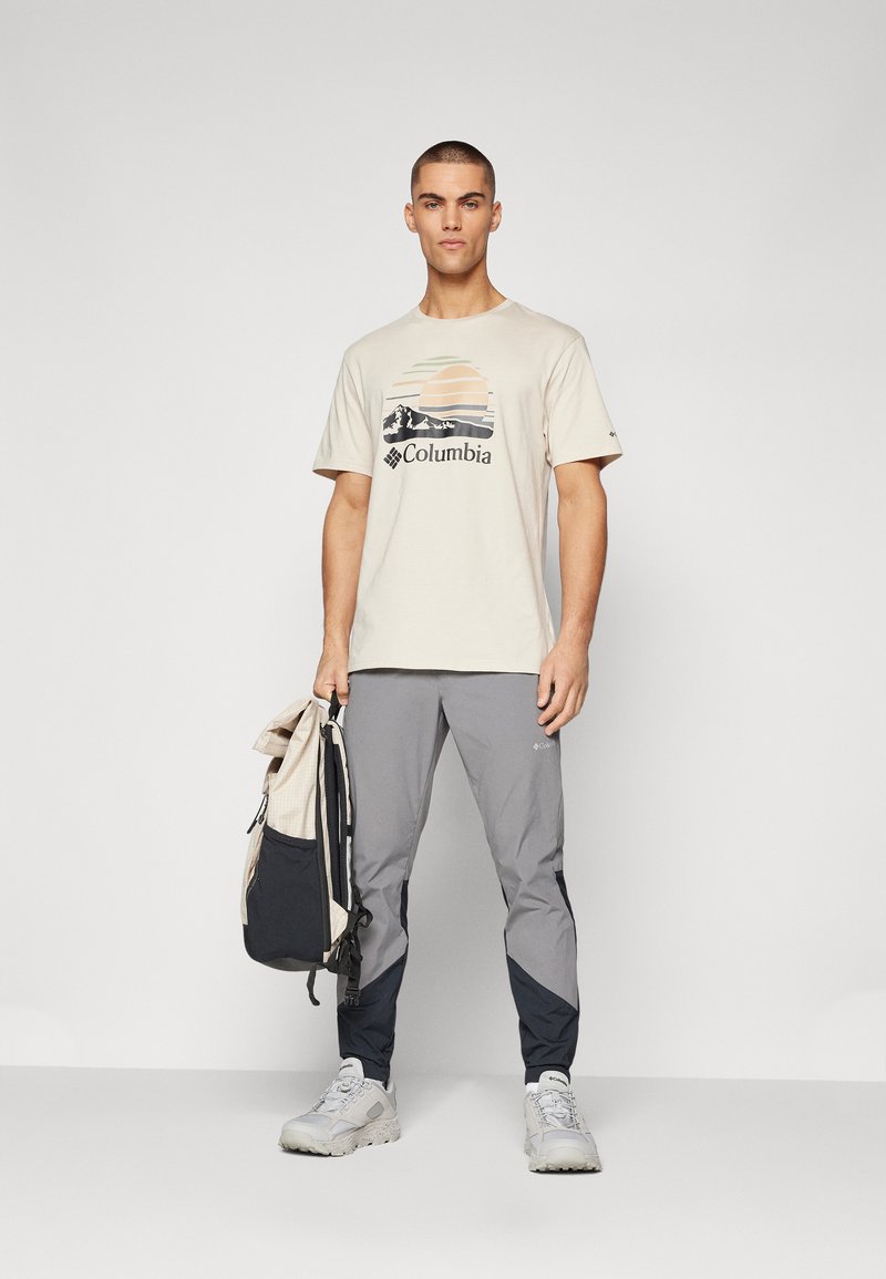Maglietta beige chiaro con un design grafico che rappresenta montagne e un tramonto, abbinata a pantaloni sportivi grigi e blu scuro e scarpe da ginnastica grigie.