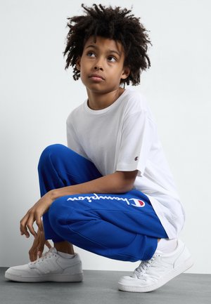 White short-sleeve t-shirt paired with blue fleece joggers featuring a logo, complemented by white sneakers. Modern sporty design and casual fit.
