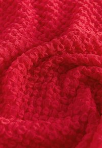 Crveni tkanina s teksturom i bogatim, istaknutim uzorkom. Materijal ima labavu tkanje, što stvara mekani, udobni izgled.