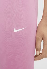 Ružové športové legíny vyrobené z hladkej, elastickej látky, s malým bielym logom Nike na ľavej strane. Detailný záber na textúru látky a strih.