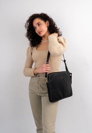 Zwarte leren schoudertas met een gladde textuur, vierkante vorm, gouden ritsen en een verstelbare strap. Draagt bij een beige trui en lichte broeken.