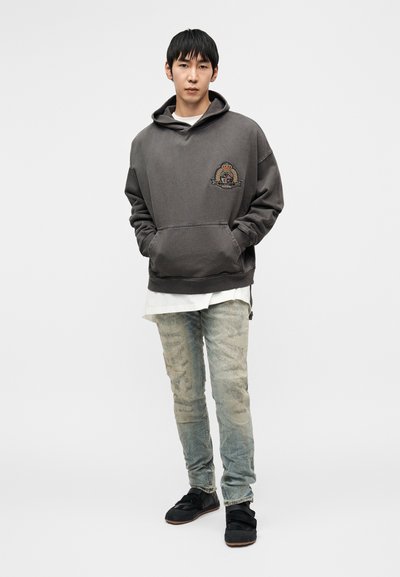 Γκρι βαμβακερή μπλούζα με κουκούλα και μπροστινή τσέπη, με κεντημένα λογότυπα. Συνδυασμένη με ανοιχτόχρωμο τζιν και μαύρα αθλητικά παπούτσια.